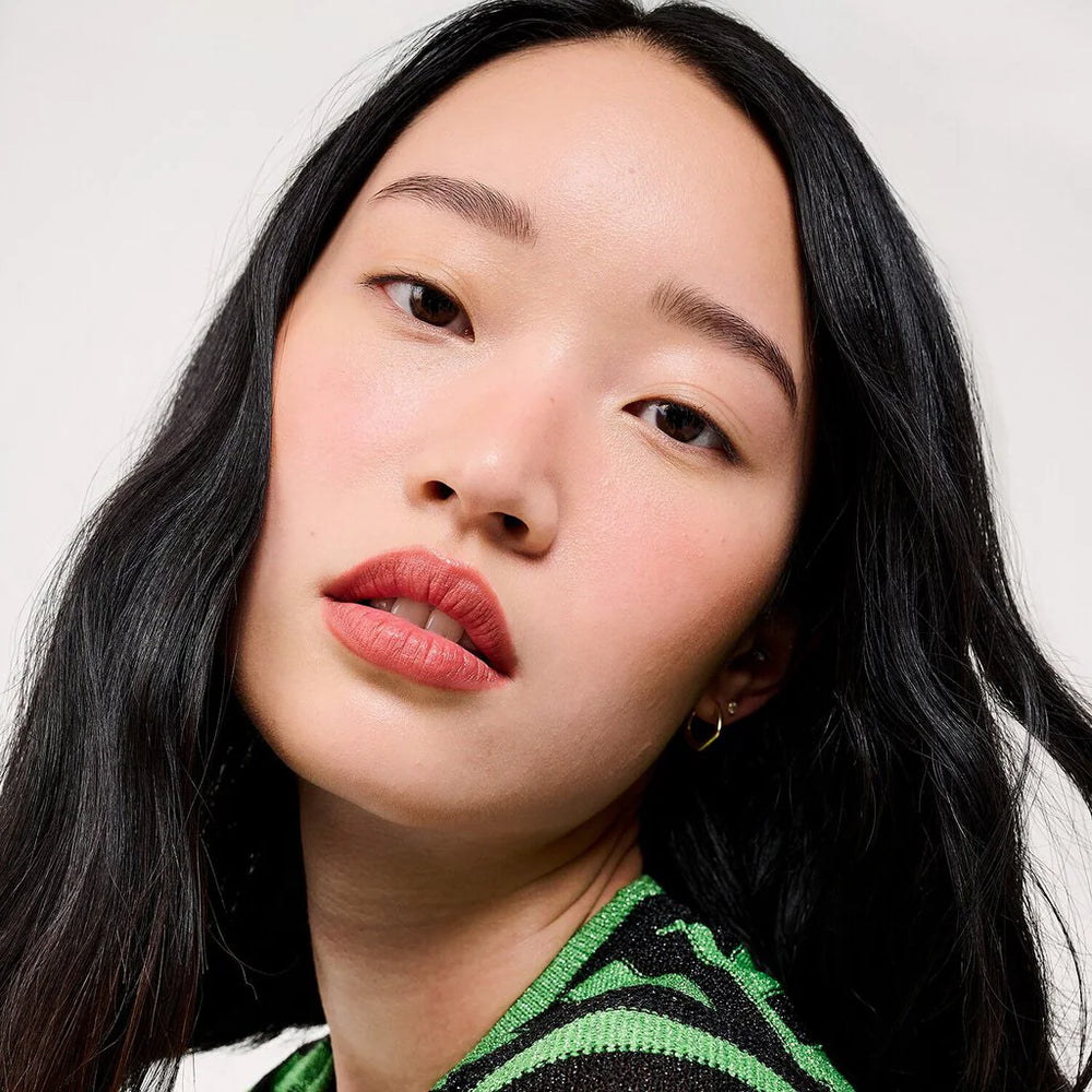 Close-up portrait of a woman with long dark hair wearing a green and black striped knit top against a light gray , Benefit The POREfessional Super Setter Setting Spraybackground.