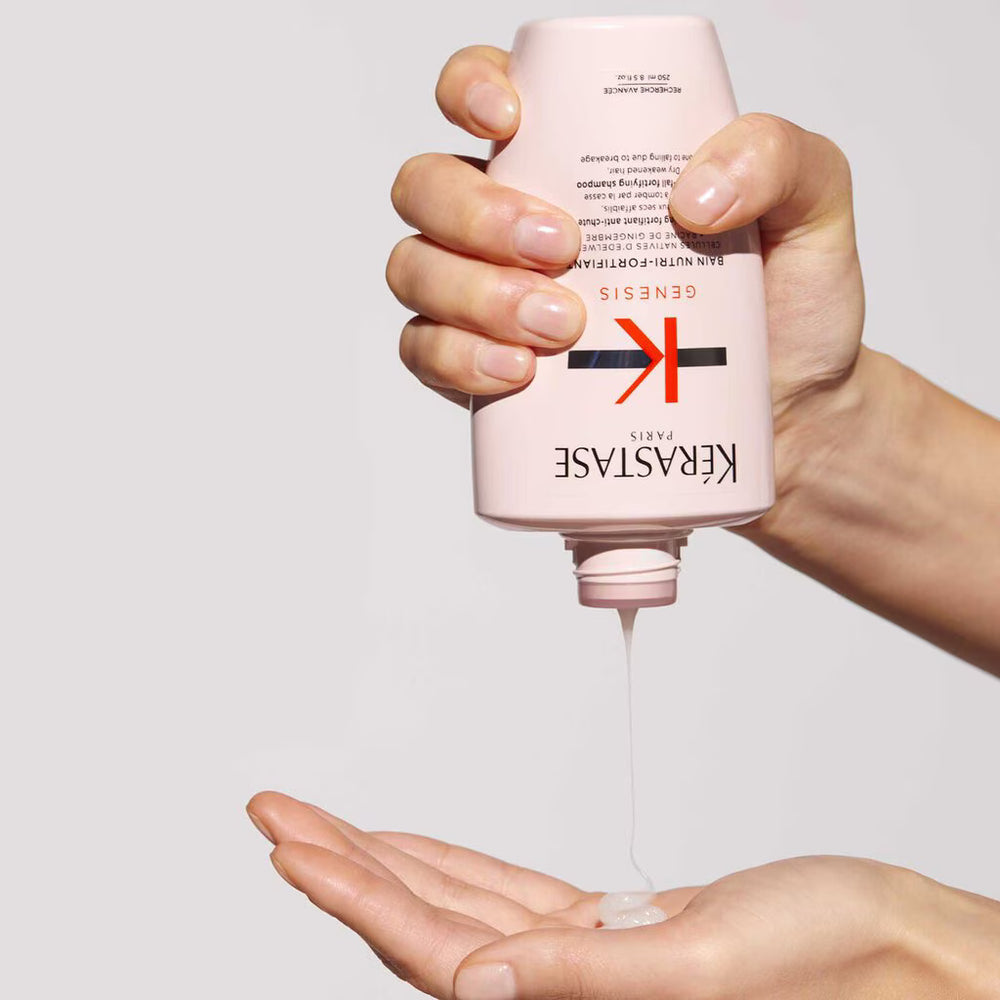 A hand holding a pink Kérastase Genesis hair developer bottle, with a stream of clear gel-like product being squeezed into an open palm against a light gray background.