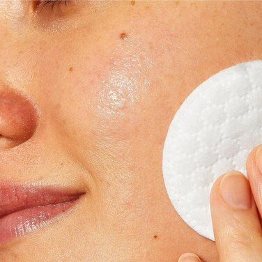 A close-up of a person's face with a white round cotton pad being pressed onto the cheek.CERAVE MICELLAIR 295ML)