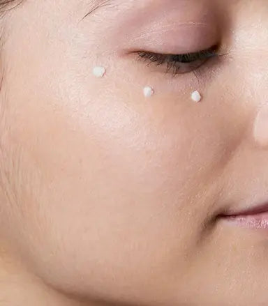 Close-up of a person's face with three small dollops of white cream applied to the cheek above the eye.Cerave Eye Repair Cream, 14ml