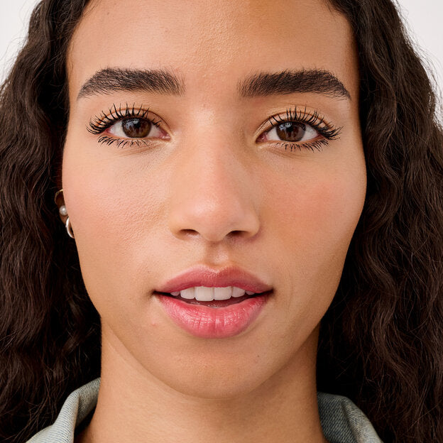 Close-up of a woman with long, dark, wavy hair wearing a light sage green top. The shot is tightly cropped to show her face, neck, and shoulders against a plain light background.benefit Waterproof Volumizing Mascara